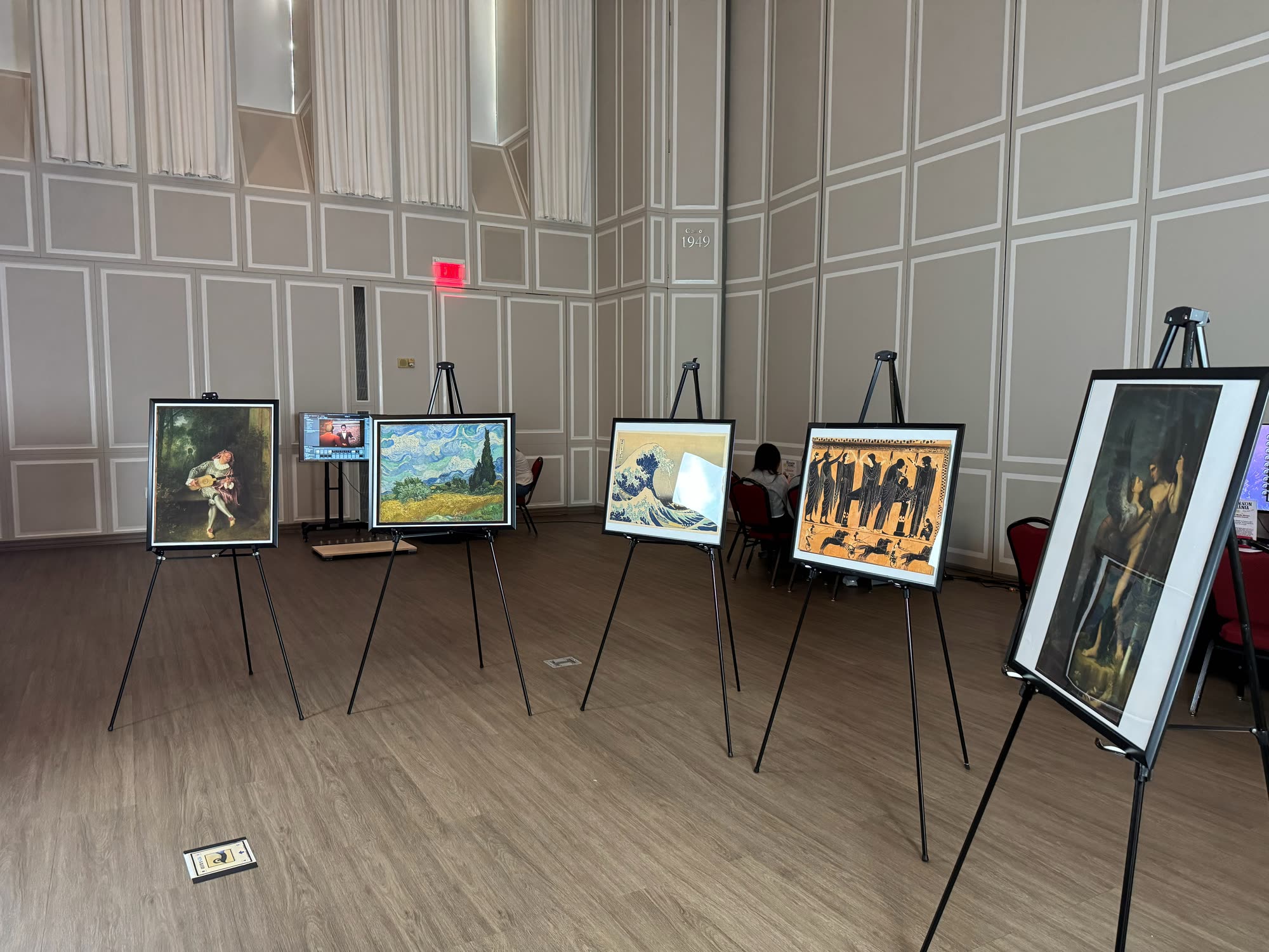 The five printed artworks on easels in a row, with a small floor marker in front.