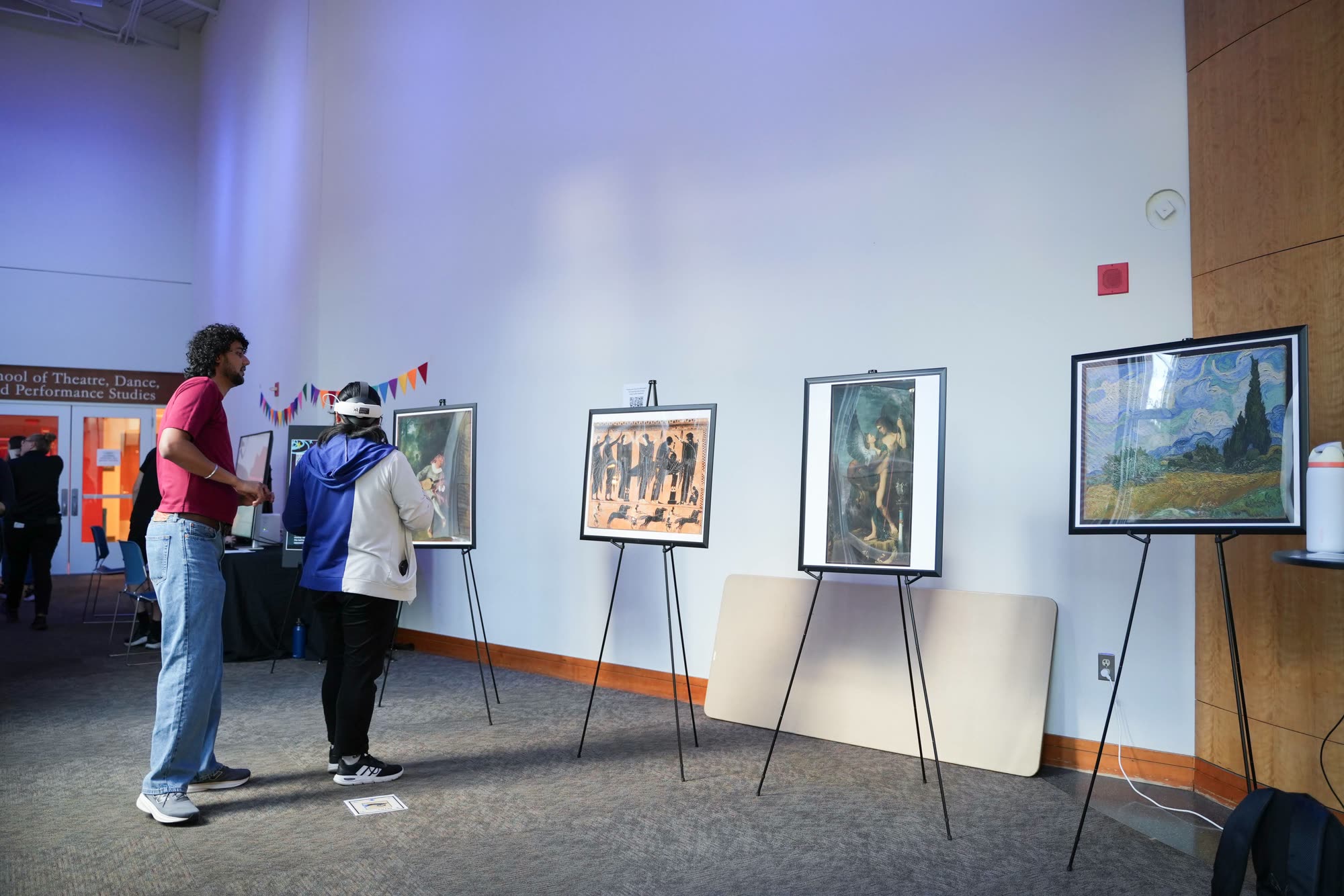Arindam guides a visitor wearing a Quest 3 headset past framed prints of the five artworks on easels.